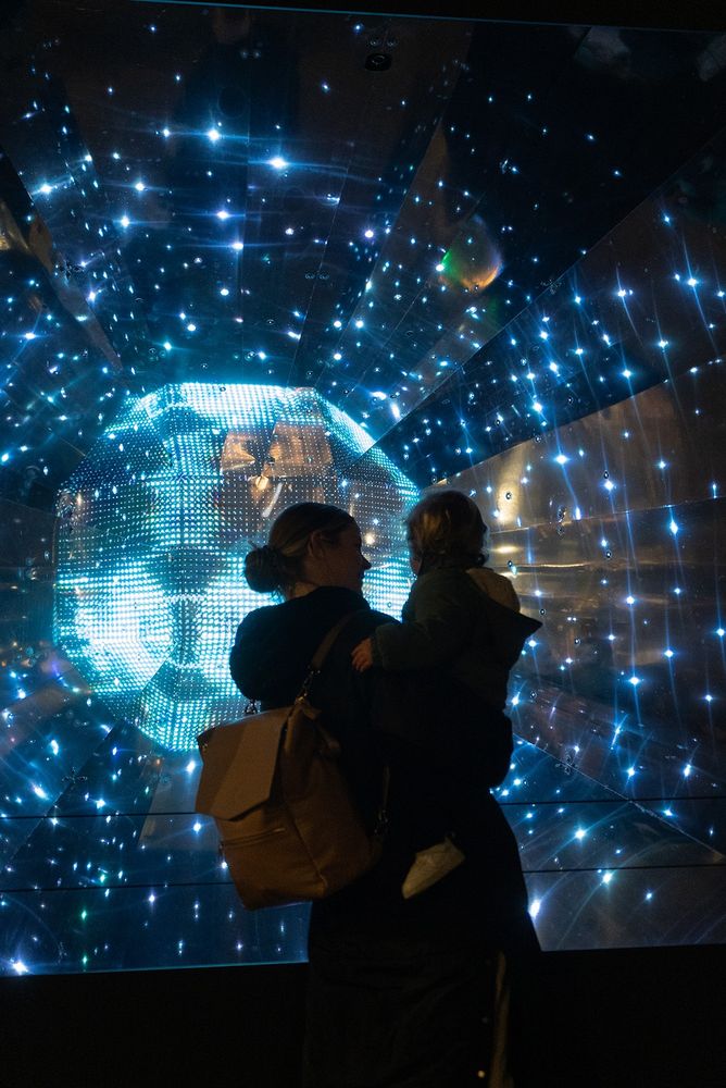 A woman holding a baby in silhouette stands in front of Geist, a large glowing orb surrounded by lights...