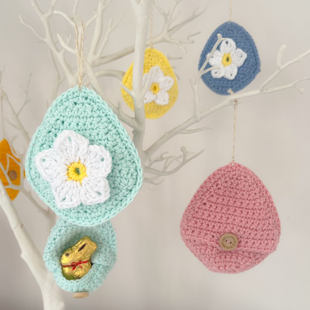 A selection of hanging crochet Easter eggs on a white twig tree. The eggs are in a variety of pastel colours, have a white crochet flower on the front and a chocolate bunny is poking out of one. 