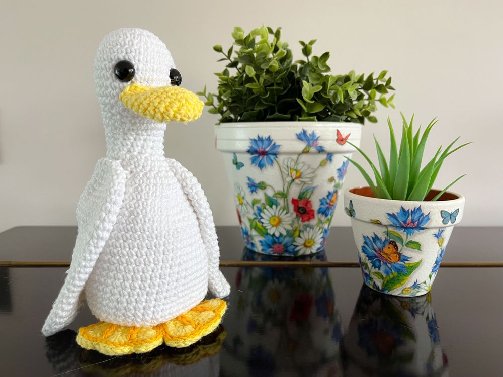 ALT: A white crochet duck stands on a black wooden top. She has been made in a crisp cotton yarn giving a clean, modern finish. She is around the same height as the larger of the plants in the background. The plant pots feature bright wild flower designs. The duck as a soft yellow beak and feet, which peak out from under the body. The feet have dark yellow detailing to give a webbed effect. Her large wings are pointed down and skim the surface.