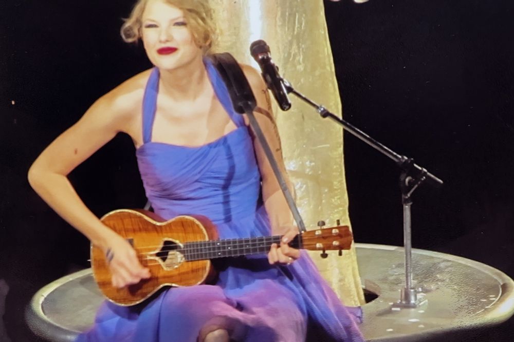 taylor swift at speak now world tour, sitting at a tree prop in the purple dress playing a ukulele and smiling