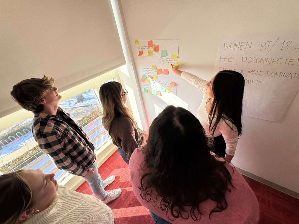 Students work with Feli Carrique of the News Product Alliance - standing in front of a white wall wiht posterboard and post-its in a brainstorming session.