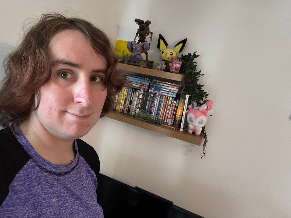 Ruby smiles slightly, a shelf full of DVDs behind her, alongside some plushies, a plant draping leaves down and a model of the Revenant from Doom