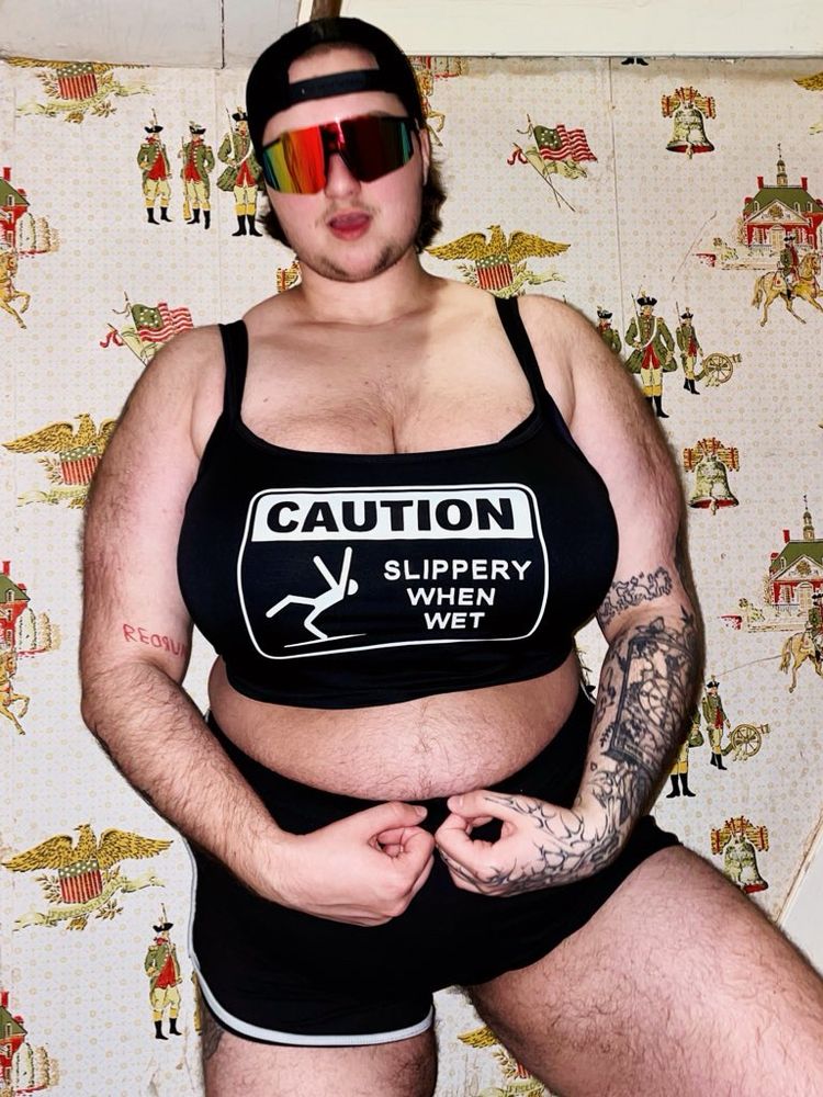 danny is flexing in a crop top that says “caution: slippery when wet” and short shorts. His hairy thighs, tummy, chest, and arms are all visible. The outfit and his cleavage looks very out of place. 