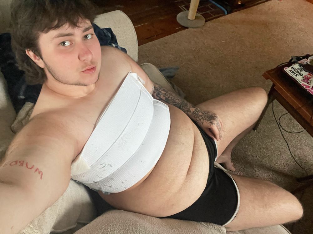 Danny is sitting on the couch with just a medical binder and black shorts on. his hairy belly is out and he’s looking up at the camera epic style 