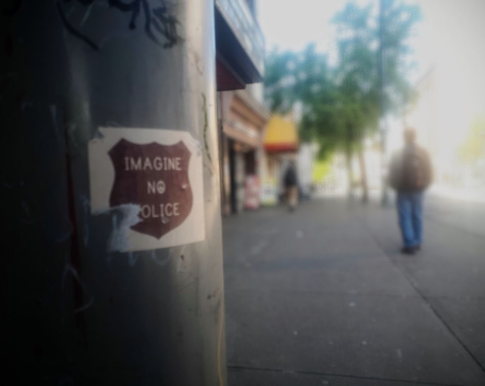 close-up of a white sticker on a pole. theres a monochrome brown badge shape, text  in white (just centred) 

IMAGINE
NO
POLICE 

the sidewalk in the background is blurred out. the sky is bright. one figure is walking away.