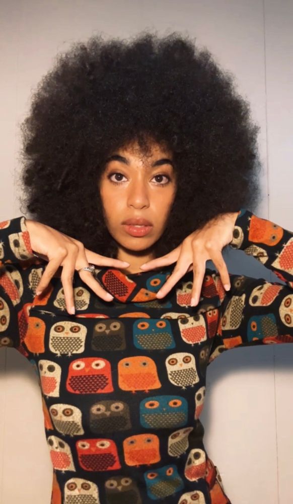 I am standing in front of an off white/egg shell wall. I am wearing an amazing thrift find: an owl dress. the owls are in various fall style colors (dark teal, off white/egg shell, bright orange, deep orange, and brown/dark grey. They are very cute. The dress has longs sleeves. My hands are in a dramatic pose, almost fanned out, framing my face. I am looking into the camera, lips parted, Afro huge.