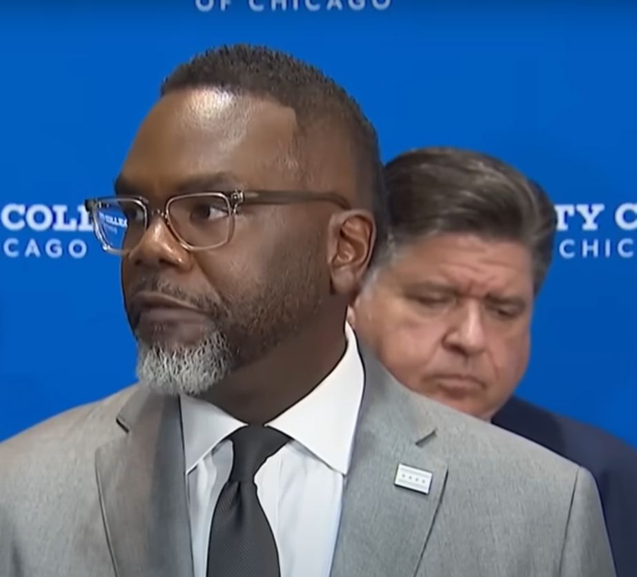 Chicago Mayor Brandon Johnson, a Black man with short black hair and a graying goatee is wearing glasses, a light suit, dark tie, and a City of Chicago lapel pin as he speaks to the press. Standing behind him, looking solemn as he glances down, is Illinois Governor JB Pritzker, a white American Jew of Ukrainian descent. He has dark hair that is graying at the temples and is wearing a dark blue suit. They are standing before a blue background on which the words "City Colleges of Chicago" appear repeatedly.