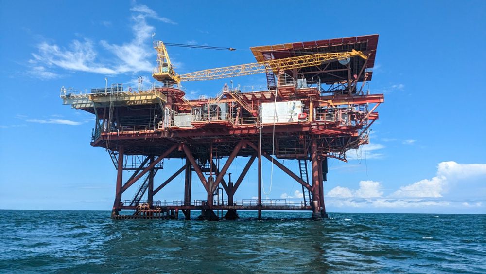 Photo of an oil rig in the Gulf of Mexico, taken from water level (in a small boat). Blue sky with a few puffy white clouds in the background. Rig is mostly red & safety yellow, standing on a few steel legs painted red. Most of the rig is visibly sun-bleached and/or covered in barnacles, visibly aged & somewhat out of use but still sturdy.