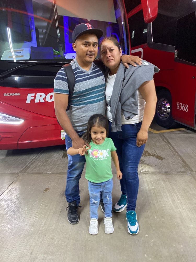 Family photo of Marco Antonio Hernández Guevara with his wife and youngest four-year-old daughter, as he was leaving for the US to work on farms. They're standing in front of coach buses at a bus station, presumably the charter buses that farm labor contractors rent to bring workers to the US.
