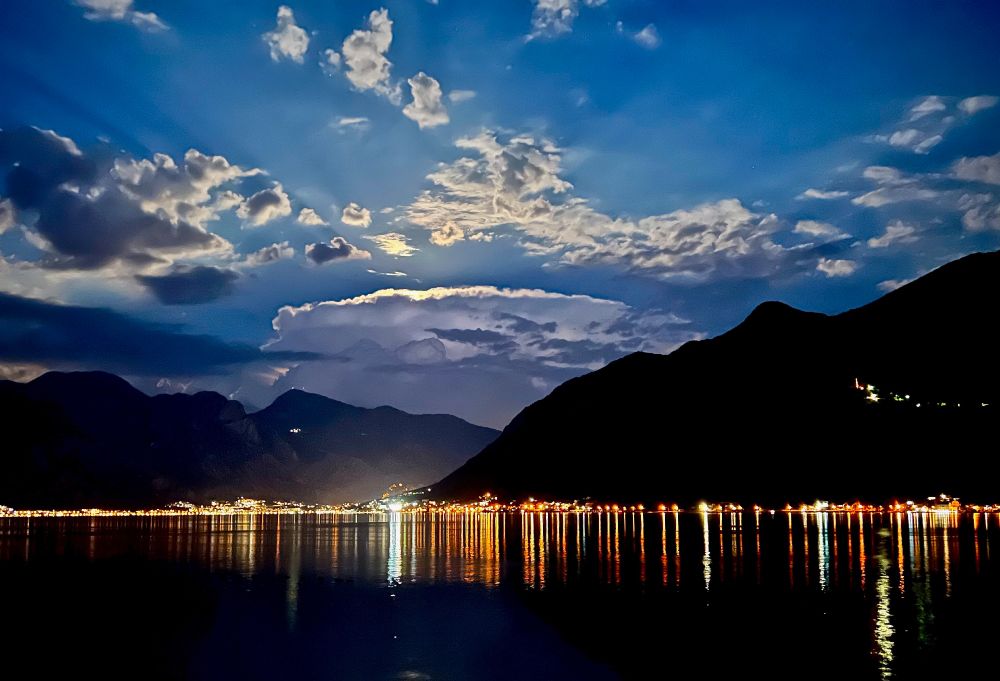 Nighttime land and seascape showing still dark water in the foreground reflecting street and houselights, as well as dark mountains in the background, under a sky filled with clouds and moonlight.