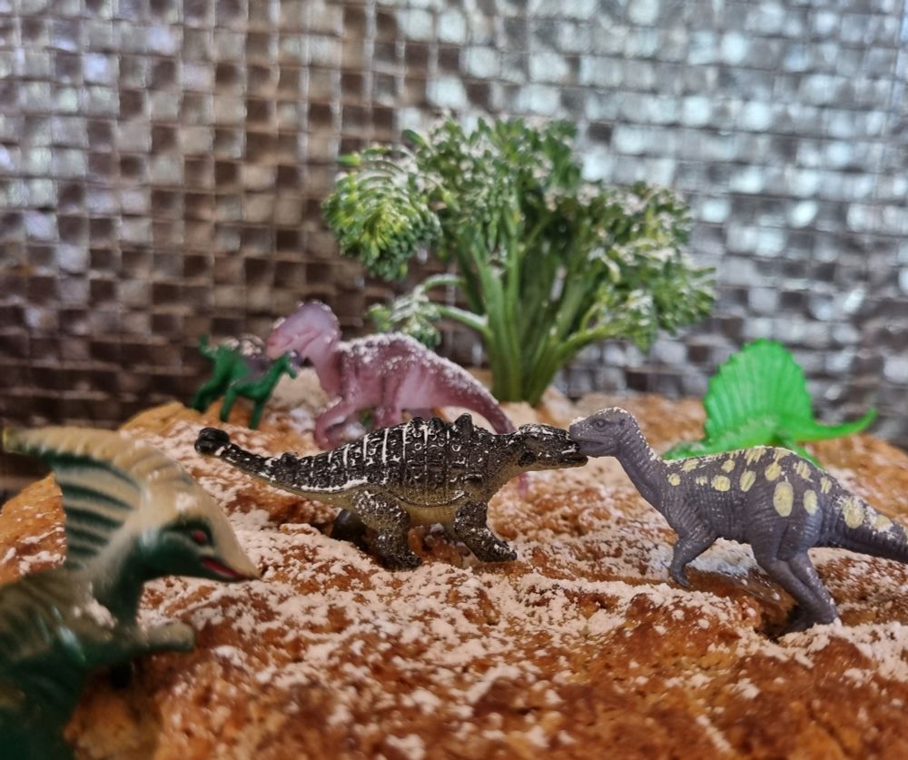 Close-up photo of a vegan coconut and lemon cake, decorated with small dinosaur toys. The crust of the cake looks like a rocky landscape. There is a broccoli tree on the cake and a dusting of icing sugar snow. 
There are dinosaurs interacting in the snow (one dinosaur is clearly getting ready for lunch, as it is biting another dinosaur's neck). 