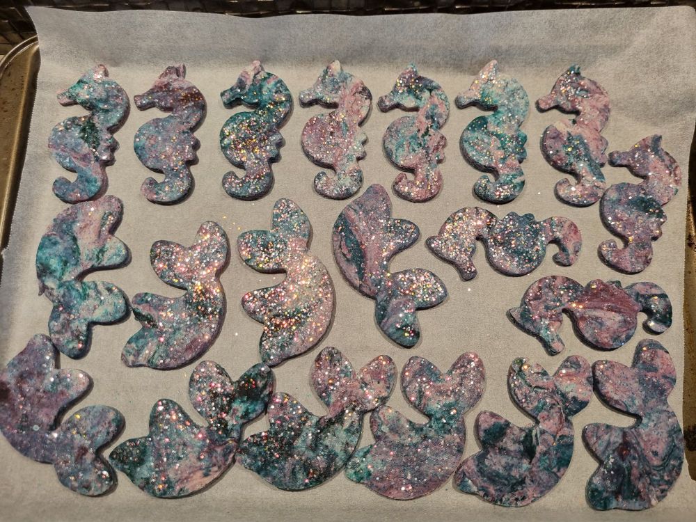 Glittery purple and teal salt dough seahorses and mermaid tails are seen on a baking tray, covered in baking paper. The ornaments look a bit galaxy-like. 