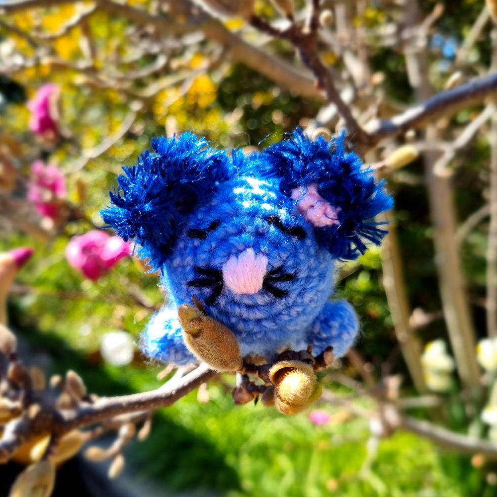 A blue possum-tiger with fluffy blue ears is sleeping happily on the branch of a flowering magnolia. It looks very content. 