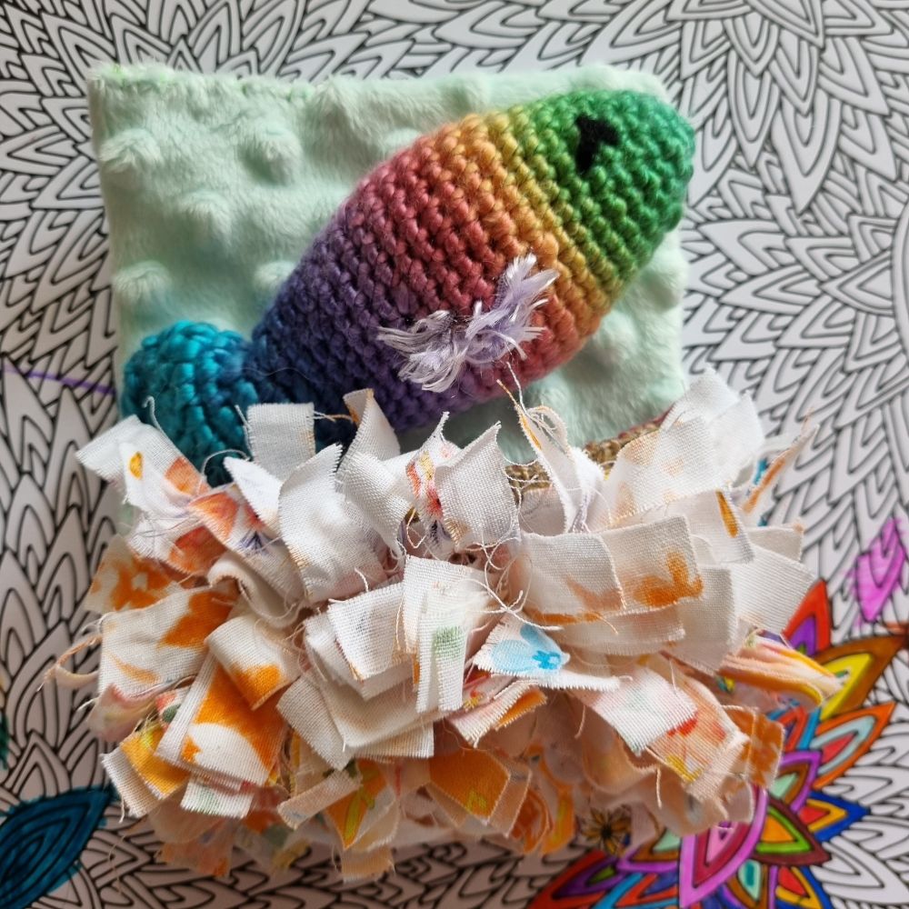 Crochet rainbow fish with fluffy purple fins, on a light green fleece mattress and under a multicoloured rag rug doona 