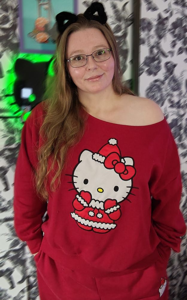 A white woman with long blonde hair, hazel eyes, glasses, cat ear hair clips, wearing a red Hello Kitty Christmas themed sweatshirt and pants. She is smiling 