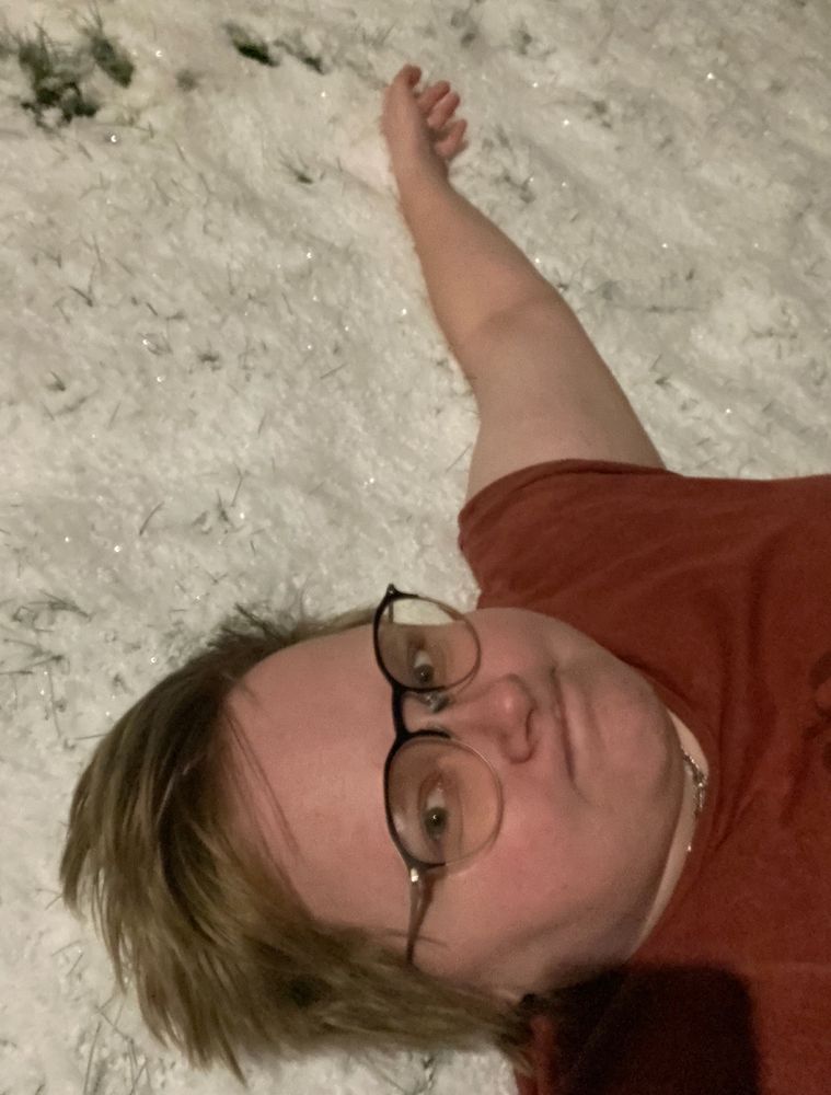 Selfie taken of alli affirmative lying in the snow in a t-shirt, you can only see shoulders up and his arm lying in the snow. His expression is a mix of stressed and unimpressed 
