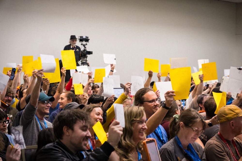 A photo of a room full of people at San Diego comic-con 2025 all holding up drawings of their octopuses. 