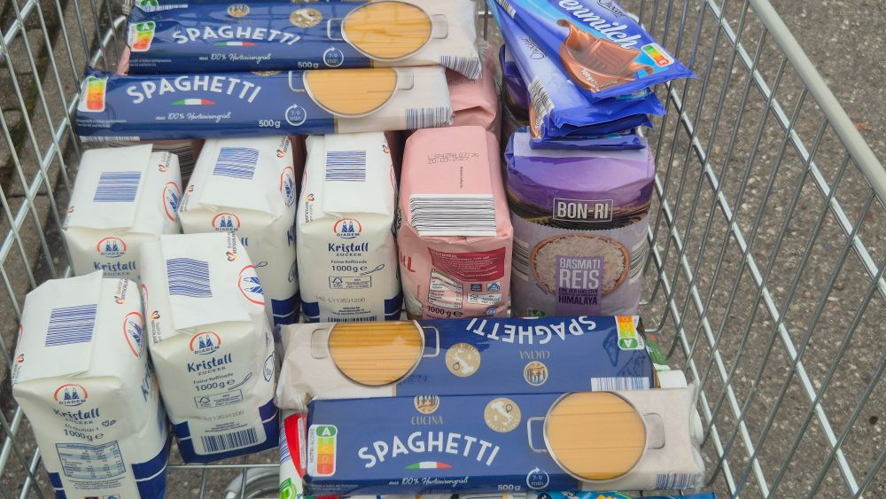 Ein Einkaufswagen gefüllt mit mehreren Packungen Nudeln, Mehl, Zucker, Schokolade, Reis...