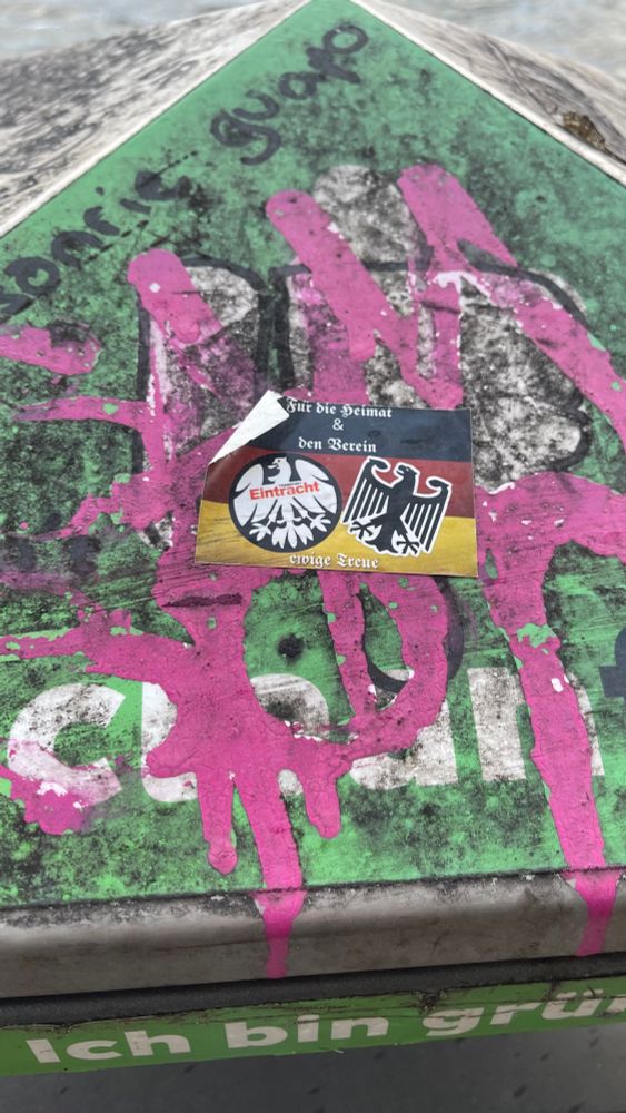 Ein Sticker mit Deutschlandflagge, der Aufschrift für die Heimat & den Verein, ewige Treue, mit dem alten Logo der Eintracht Frankfurt (schwarzer Kreis, weißer Adler, Eintracht Schriftzug in rot) & dem Bundesadler, auf einer mit Graffiti besprühten grünen Mülltonne klebend.