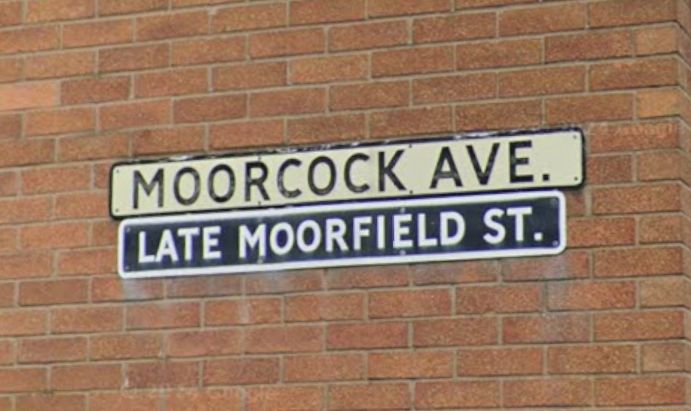 A streetname sign combo on a 60s orangey brick wall, reading "Moorcock Ave, Late Moorfield St."