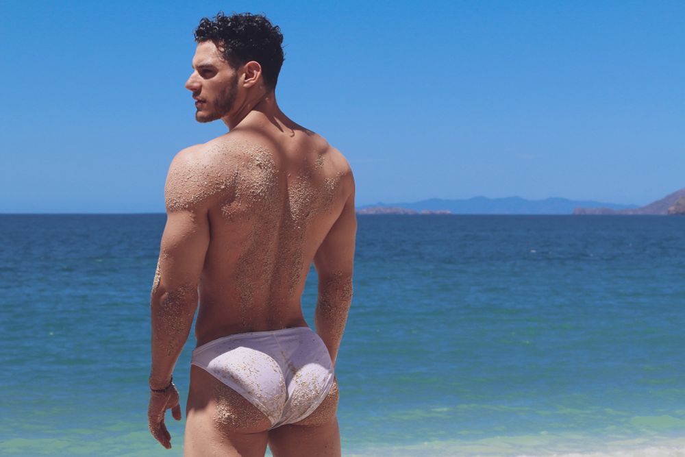 Male model posing on the beach in white swim trunks, that shows off his beautiful back. With the blueish ocean as background 