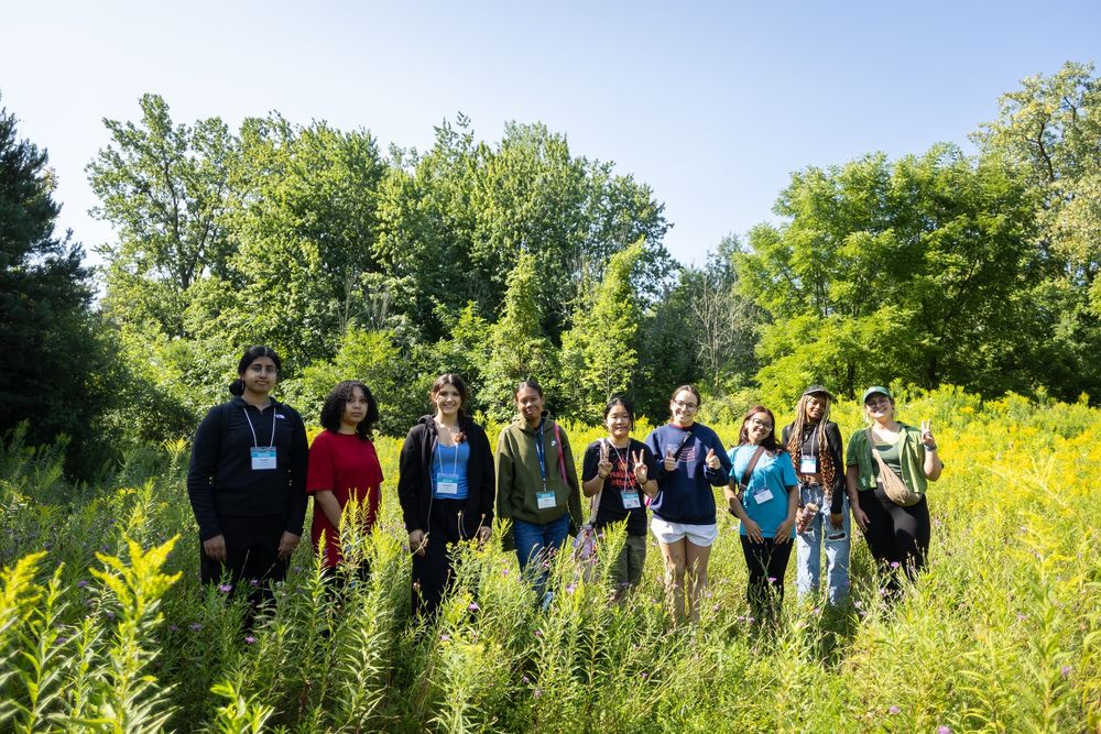 A group of nine students stands in a lush field surrounded by tall greenery and wildflowers, with trees and blue sky in the background. Most are smiling and making peace signs. They wear name badges and a mix of casual clothes including hoodies, T-shirts, and backpacks.
