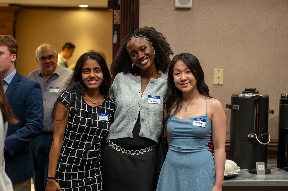 Three students pose together at an indoor reception. All are wearing nametags: one in a black-and-white checkered dress, one in a light blue sparkly dress, and one in a gray sweater and black skirt with a silver belt. They are smiling with arms around each other. A coffee station and other guests are visible behind them.