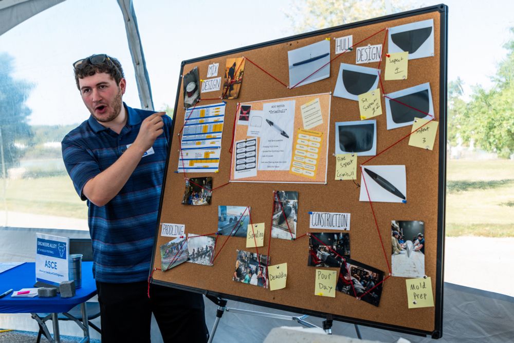 A UB ASCE student explains a corkboard display at this year's Engineers Alley at UB Football. The board has sections labeled "Mix Design," "Hull Design," "Paddling," and "Construction," connected by red string to photos and notes, including "Most Wanted: 'Crimibull.'"