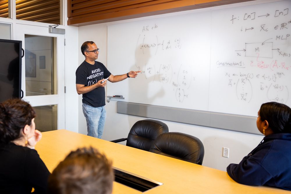 The new Department of AI and Society's inaugural chair, Atri Rudra, stands at a whiteboard, pointing to a complex math equation written in marker. He is teaching a small group of students who are attentively listening seated around a conference table.