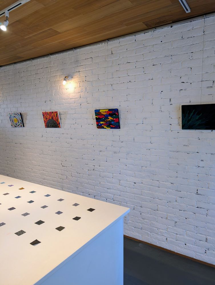 白く塗装されたレンガの壁に、4枚のカラフルな抽象的な絵画がワイヤーで吊るされて展示されています。手前には、小さな正方形の作品が並べられた白い展示台の一部が写り込んでいます。