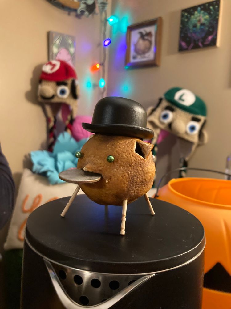 A photo of a lemon made to look like a pig with pins for eyes and toothpick legs. He is wearing a little bowler hat. 