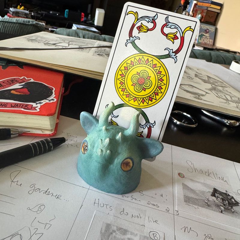 A messy artist desk with sketches and notebooks scattered about. In the middle of the photo is the head of a blue creature with horns and yellow eyes, holding a tarot card (Ace of coins, Marseille deck) 
