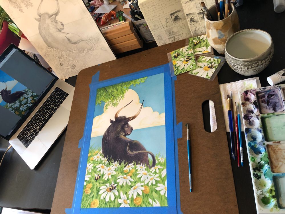 Messy artists table: from left to right: an open laptop, a drawing of a bull on white paper, misc art supplies and piles of brushes, a big palette of paint. In the center is a wooden board with blue tape holding down a painting of a bull in a field of daisies. 