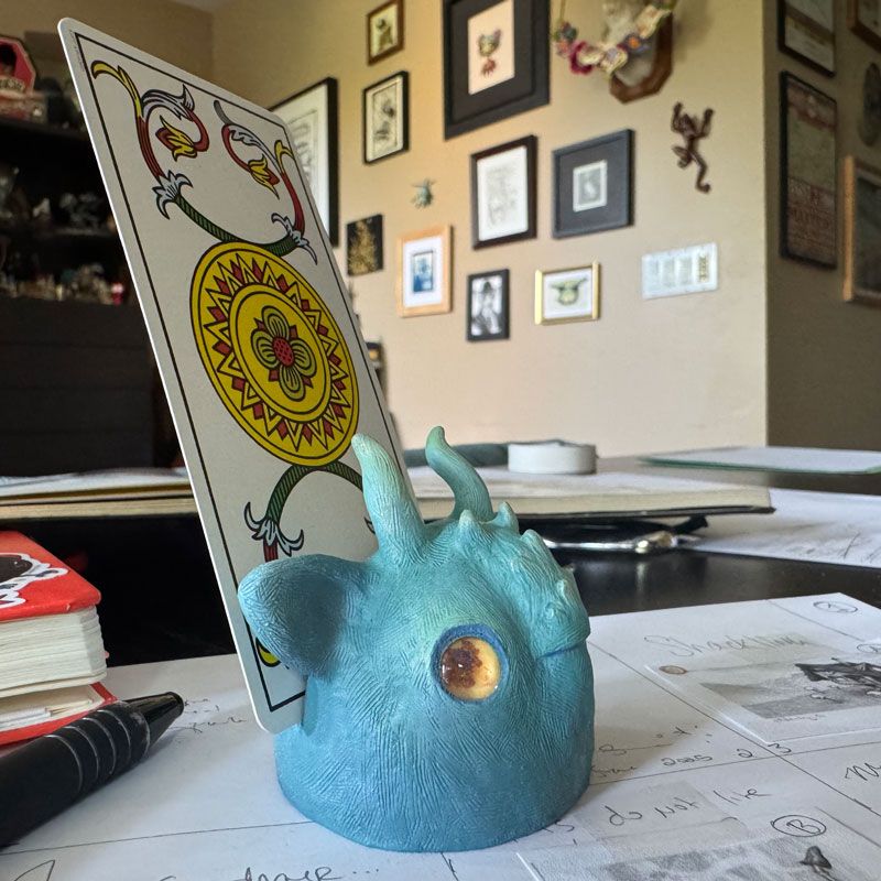 A messy artist desk with sketches and notebooks scattered about. In the middle of the photo is the head of a blue creature with horns and yellow eyes, holding a tarot card (Ace of coins, Marseille deck) 