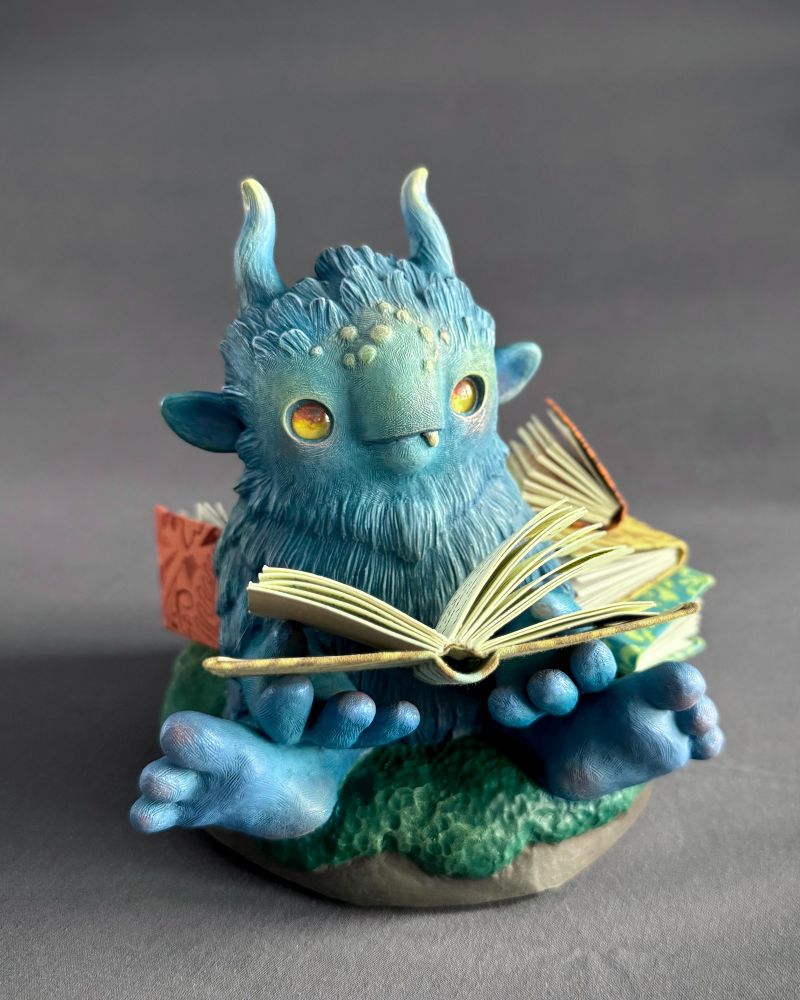 A clay sculpture of a blue, hairy beast with golden yellow eyes and blue horns. It's holding a book with some other books in the background.