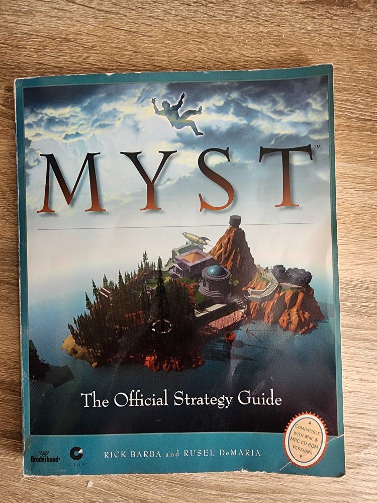 The front cover of "Myst, The Official Strategy Guide" by Rick Barba and Rusel DeMaria. It's a blue paperback book with a large landscape view of the Myst Island, all the unique areas of the island visible.