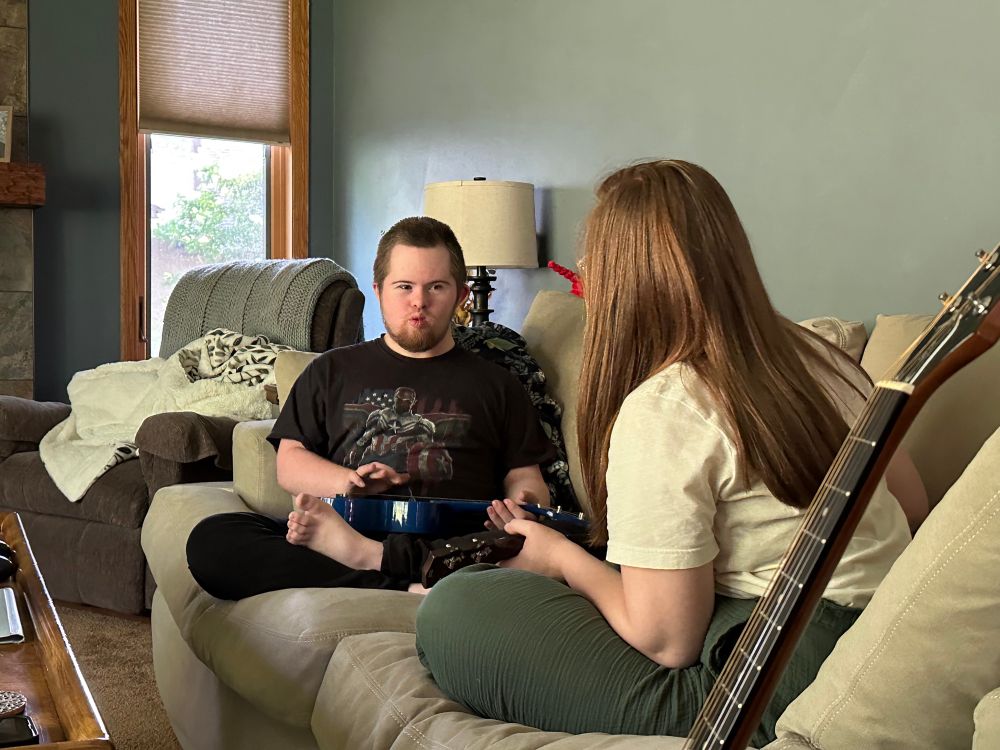A young man with Down Syndrome sits on a couch with a ukulele in his lap. A young woman with red hair and her back to the camera faces him on the couch. 