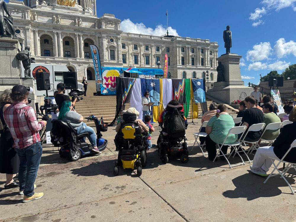 A group of people sit and stand to listen to Peggy Flanagan in front of the Capitol. 