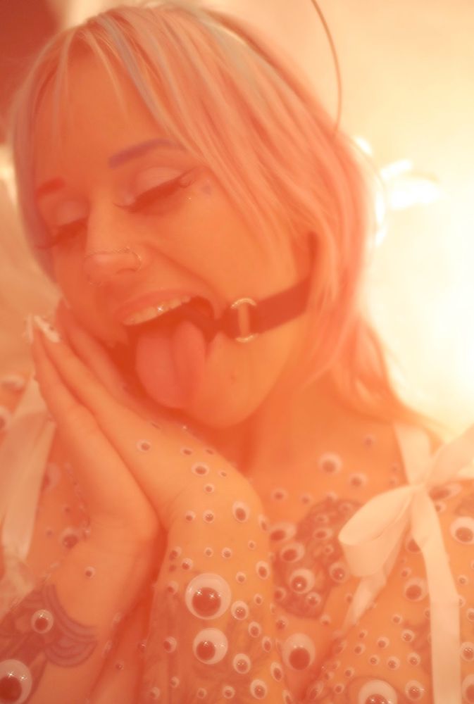 AFAB sits in white lingerie with their tongue sticking out of a ring gag on. their body is covered in googly eyes and their hands are pressed together with their cheek resting on them. they are adorned with a halo and angel wings. 