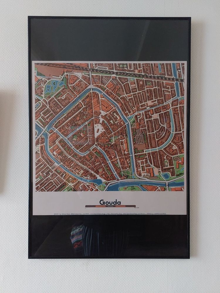 Print van 60 bij 60 cm on een zwarte lijst met een zwarte passe-partout, met als afbeelding een plattegrond van de binnenstad van Gpuda, in de kleurenoranje, beige, blauw en groen
