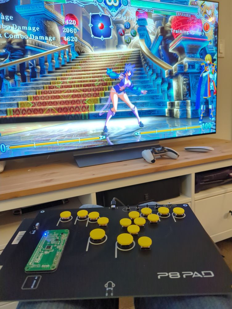 View of a custom PCB lever-less fighting game controller on a lap. The controller is plugged into a PS5 and shows BlazBlue Central Fiction on the TV with the character Bullet in the training mode.
