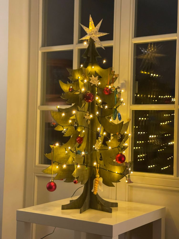 Zu sehen ist ein geschmückter und beleuchteter kleiner Weihnachtsbaum, der auf einem weißen Tisch steht und sich im dahinter befindlichen Fenster spiegelt. 

Der Weihnachtsbaum ist aus Holzplatten gefertigt, die vertikal in einander gesteckt werden. Es ist eine Laubsägearbeit und grün lasiert. 
