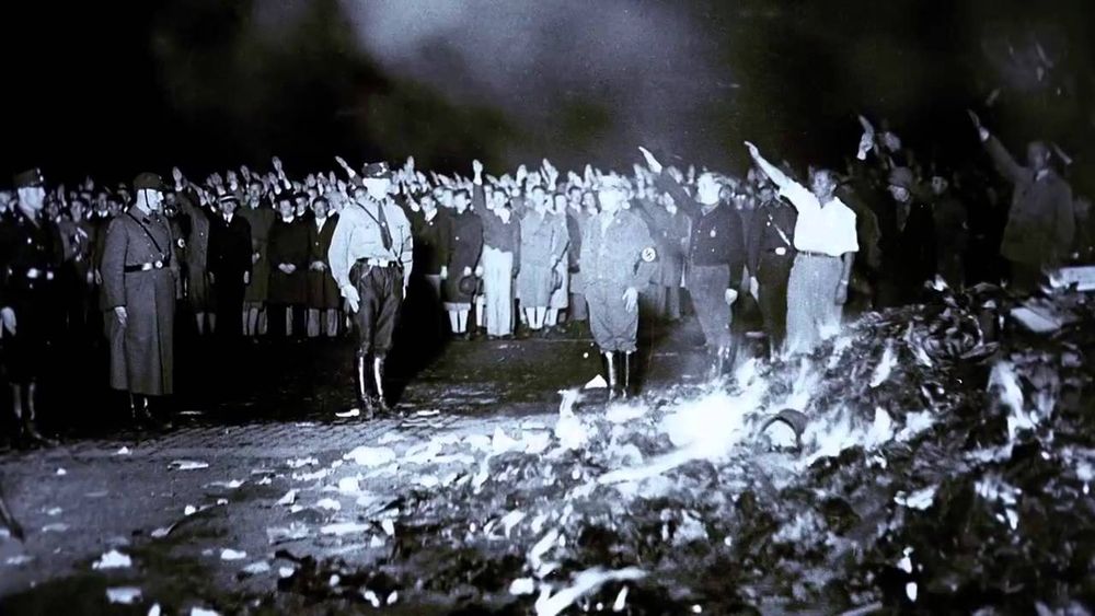 Black and white image depicting a sizeable group of Nazis in uniforms and civilians standing in the streets next to a large pile of burnt and burning books. Image taken May 10th, 1933 in Berlin, Germany.
