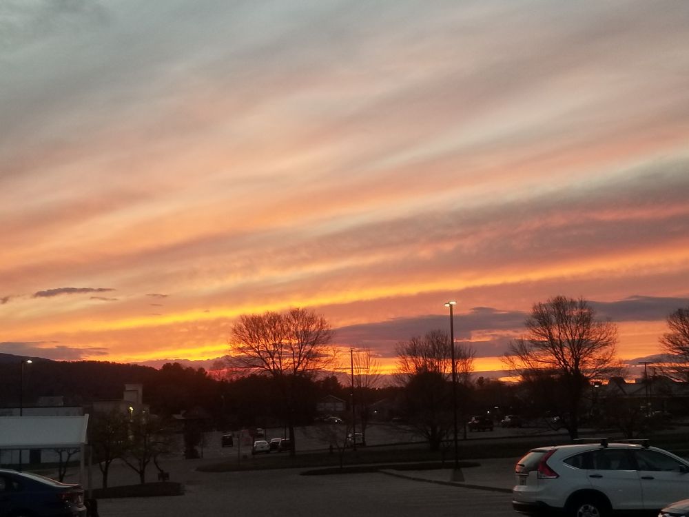 Orange/red sunset with parking lot and strip mall in the foreground. Small number of parked cars. Trees with no leaves. 