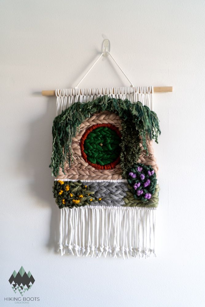 A woven tapestry hanging in a wall. It features a Lord of the Rings inspired Hobbit home with a round door, flower bushes and ivy/greenery draping over the top of the house. It is made of woven wool and yarn.
