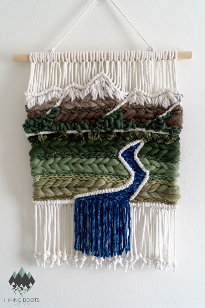 A woven tapestry that uses wool and yarn to create a fluffy and textured mountain scene. It has a range of snow-capped mountains, rows of evergreen trees, and a meadow with a blue stream running through it.