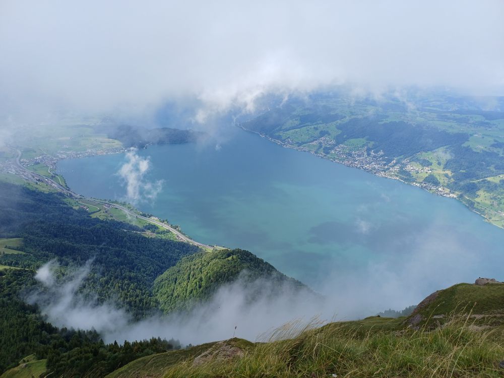The view of a mountain top: you see a lake, a lot of clouds and a green landscape.