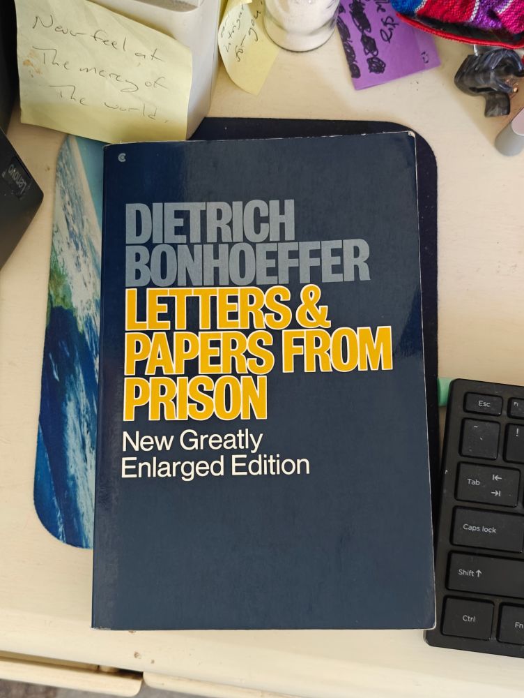 A book, Letters & Papers From Prison by Dietrich Bonhoeffer, sits on a desk. In the upper left corner of the photograph is a post-it note that reads: Never feel at the mercy of the world.
