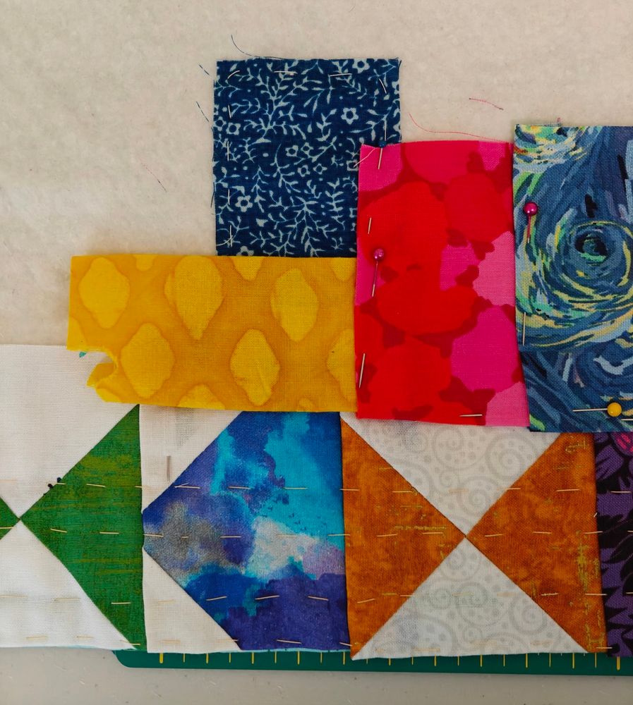 Hourglass blocks and a house block edge a quilt perimeter with yellow, red and blue scraps pinned in place above them. 