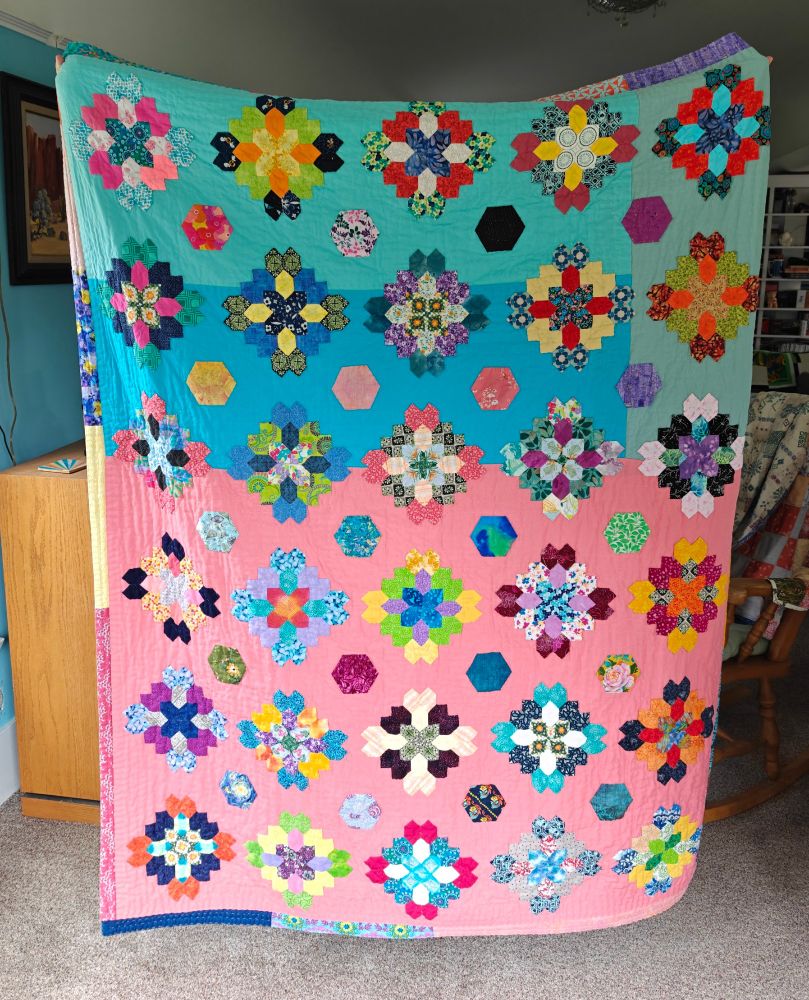 Large quilt held aloft in a living room. The quilt is decorated with on-point diamond-shaped English paper pieced blocks in the Lucy Boston pattern, all made from honeycomb shapes. Interspersed within the thirty blocks, all made from scrap fabric, are two-inch hexagons in a variety of prints. The backing fabric are three different blues forming the top third, with a block of pink as the remainder. The blocks are appliqued onto the those fabrics, bordered by strips of prints in a variety of colours. 